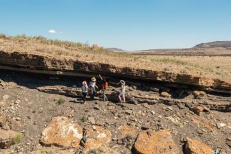 Seen from a distance, four students examine striations in red volcanic rock on the edge of a crater strewn with boulders. They wear sun hats to protect from the glare and heat. One points at the rock patterns, which are crumbled and eroded.