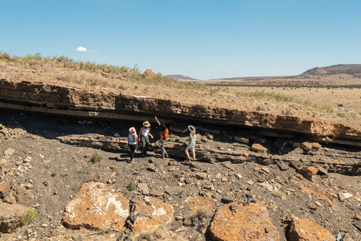 Seen from a distance, four students examine striations in red volcanic rock on the edge of a crater strewn with boulders. They wear sun hats to protect from the glare and heat. One points at the rock patterns, which are crumbled and eroded.