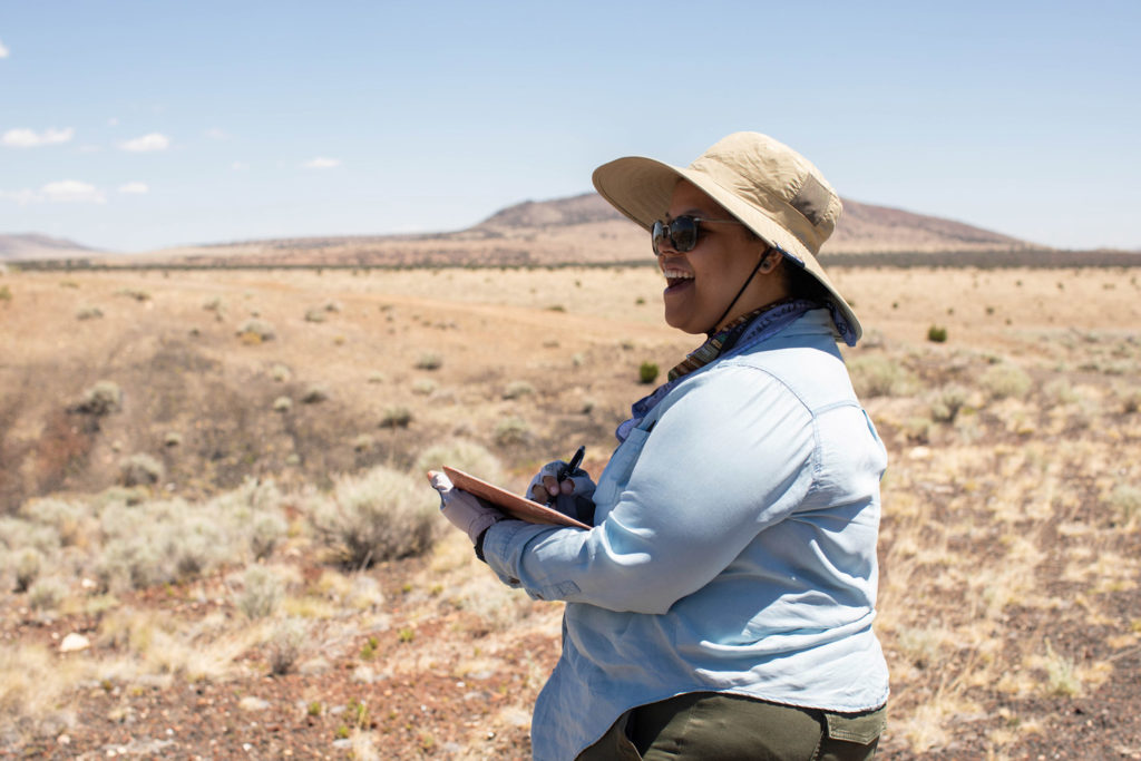 GeoSPACE program manager Yesenia Arroyo in sunglasses, a sun hat and long sleeve shirt standing in front of an extinct volcano with dry desert grasses covering the foreground. Arroyo is writing on a clipboard and smiling.
