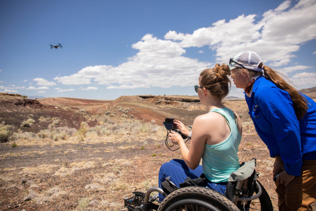 A drone flies above dry grass and rolling hills in a blue sky traced with thin white clouds. Frankie Butler, controls the drone while seated in her wheelchair, looking away from the camera toward the drone on the left. Professor Anita Marshall in a Gator blue shirt supervises, leaning down to see the screen of the controller Butler holds.