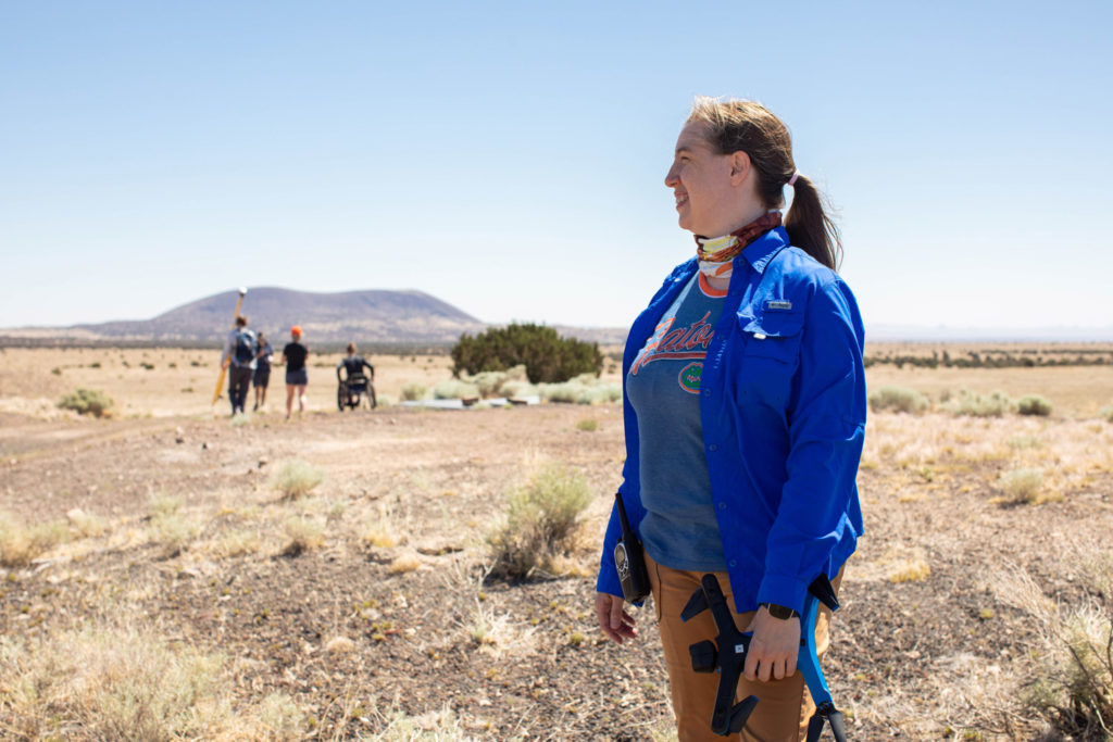 Anita Marshall, wearing a Gator shirt and holding a drone, looks out to the left over a dry grassy field with four students in the background. One uses a wheelchair, another carries a pole for GPS mapping. The sky is hazy with heat.