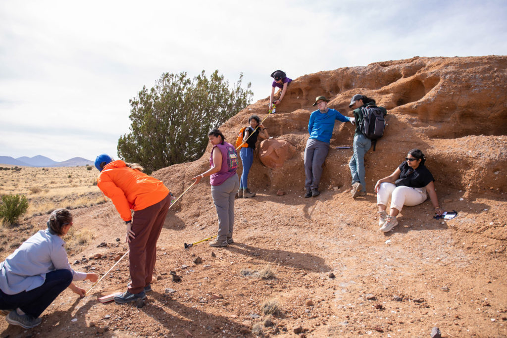 Three faculty members and five students take measurements with a measuring tape starting at a hill and going up about 15 feet to the top of a red colored rock. Some lean against the rock and sit to rest.