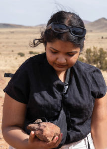 Student Shivani Dattani holds a rust-colored rock in the desert, examining it.