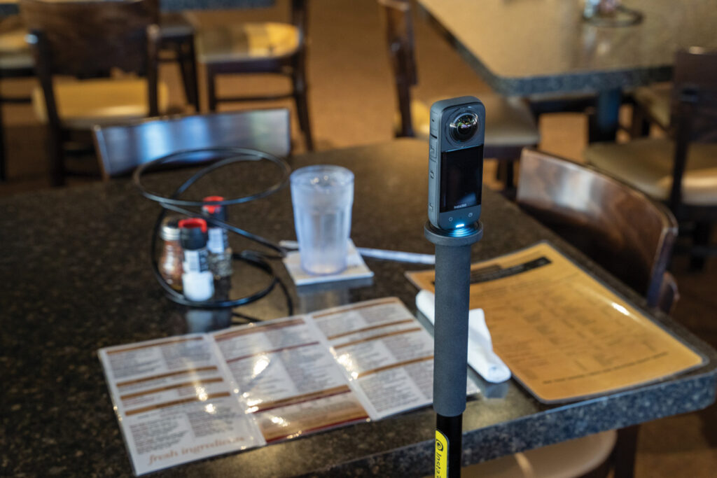 A small camera sits on top of a tripod pointed at a table in a restaurant where a menu and a glass of water sit on a table. 
