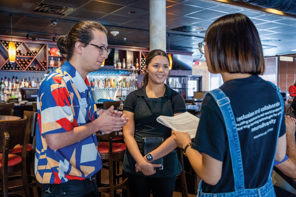 A man talks with a woman holding a script while a restaurant server holding an order pad looks on. They are in a dimly lit restaurant.
