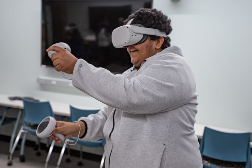 A teenage boy in a hoodie smiles as he wears a VR headset and holds two controllers. 