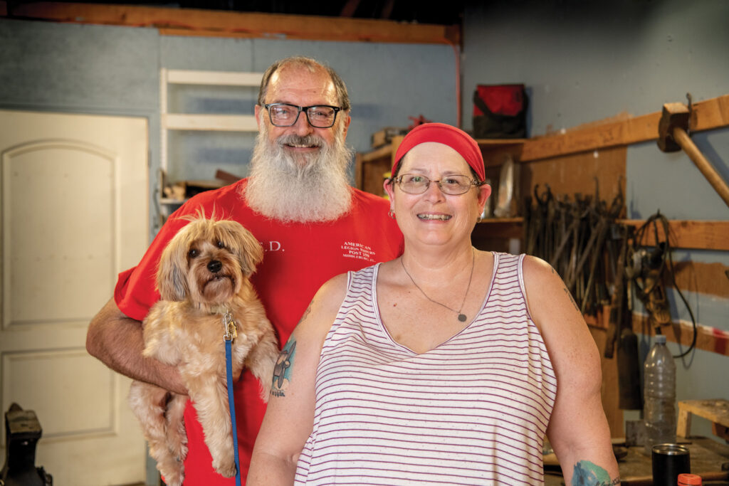 Portrait of Keith and Stephanie Early with their service dog, Riley. They are standing in the blacksmith's work shop.