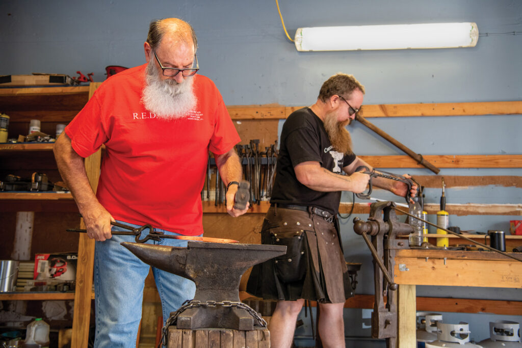 Keith Early hammers glowing metal on an anvil while the blacksmith in a kilt stands behind him at a work table. Both have long beards and protective glasses. 