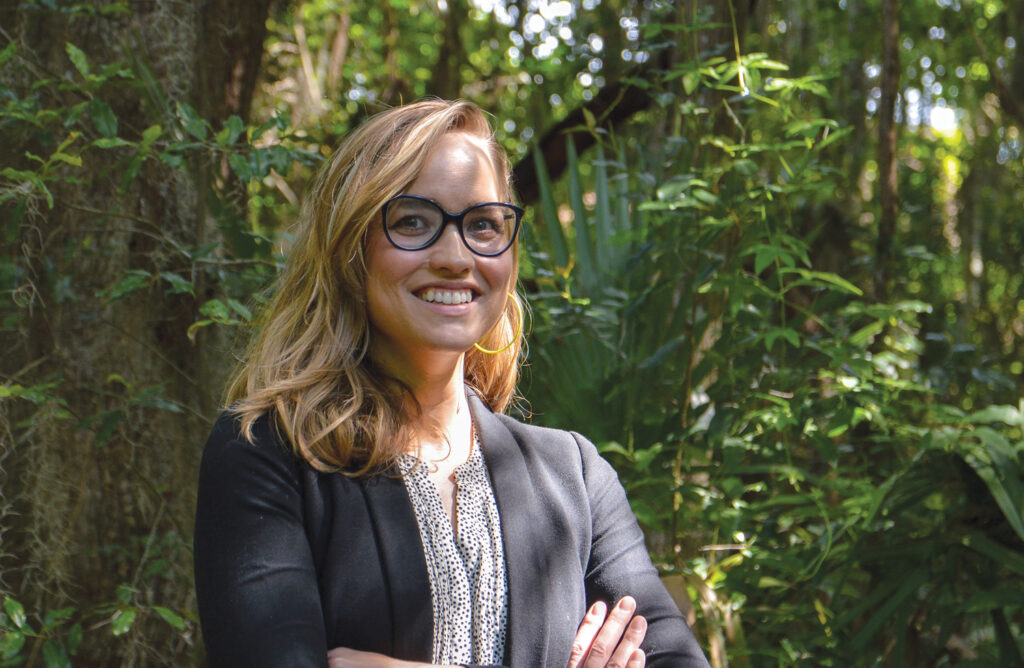 Meredith Berry stands with arms crossed in the dappled shade of a wooded area. 
