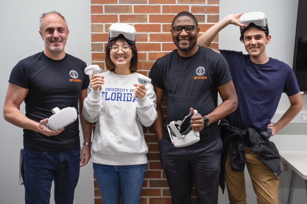 Four people stand near a brick wall in a classroom space holding virtual reality headsets and controllers while smiling. Two wear the lab logo tshirts while one wears a University of Florida sweatshirt.