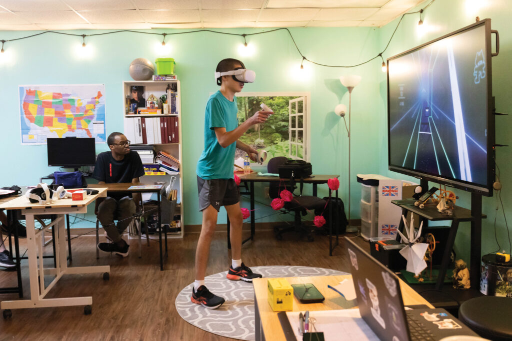 A cheerful classroom with mint green walls lit by string lights across the ceiling. A young man wears a VR controller headset and plays a game with light sabers while his classmate sits at a nearby table and watches. 