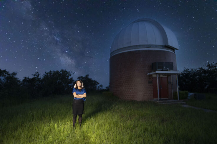 Sheila Sagear stands in front of the dark sky where the Milky Way shines as a cloudy white stripe in the sky. Behind her is UF's Rosemary Hill Observatory, a brick cylindrical building with a half-dome white roof.