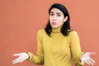 An image of a dark haired woman, wearing a yellow sweater, shrugging her shoulders.