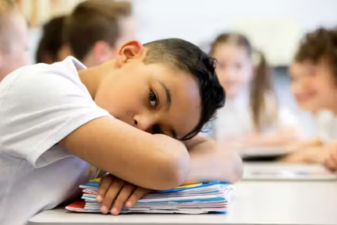 An image of a boy laying his left side of his face on his arms against a table.