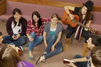 An image of eight teenagers sitting in a circle, one is holding a guitar.