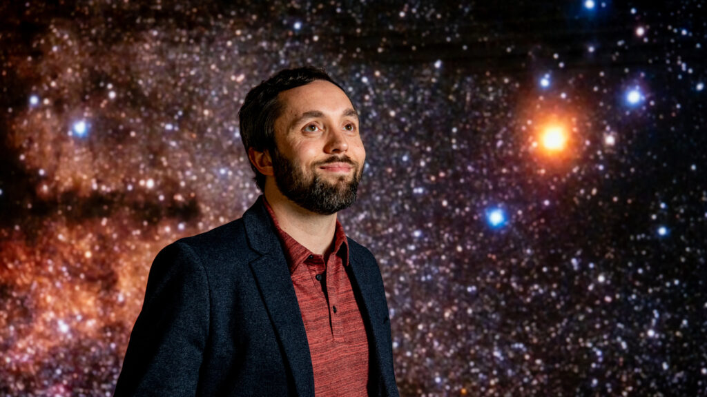 Portrait of Jason Dittmann standing in front of a screen showing a starry backdround.