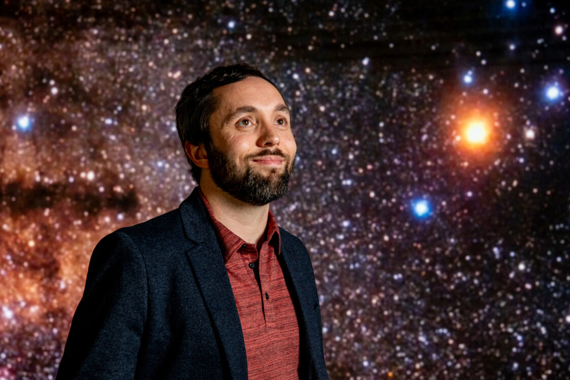 Portrait of Jason Dittmann standing in front of a screen showing a starry backdround.