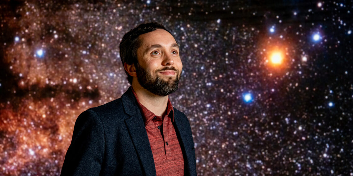 Portrait of Jason Dittmann standing in front of a screen showing a starry backdround.