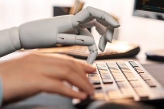 An image of human and robot hands typing on a computer keyboard