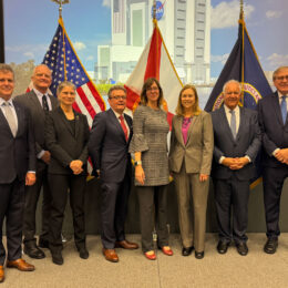 A group of eight individuals stands together in front of flags representing the United States and Florida. They are posed in a conference room with a large screen displaying a building in the background. The group appears professional, wearing business attire and smiling for the photo.