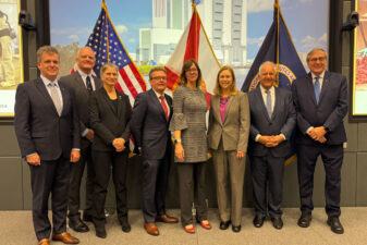 A group of eight individuals stands together in front of flags representing the United States and Florida. They are posed in a conference room with a large screen displaying a building in the background. The group appears professional, wearing business attire and smiling for the photo.