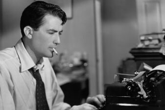 A black and white photo of a white male sitting at a typewriter with a cigarette in his mouth.