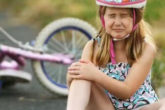 An image of a young girl holding her right arm in pain, wearing a bike helmet with her bike the background.