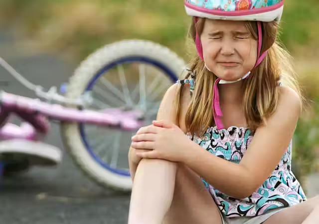 An image of a young girl holding her right arm in pain, wearing a bike helmet with her bike the background.