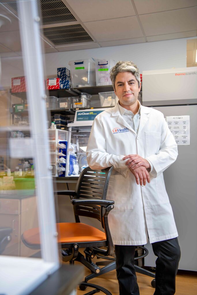 A person standing in a white lab coat, leaning on a chair with wheels with lab equipment in the background.
